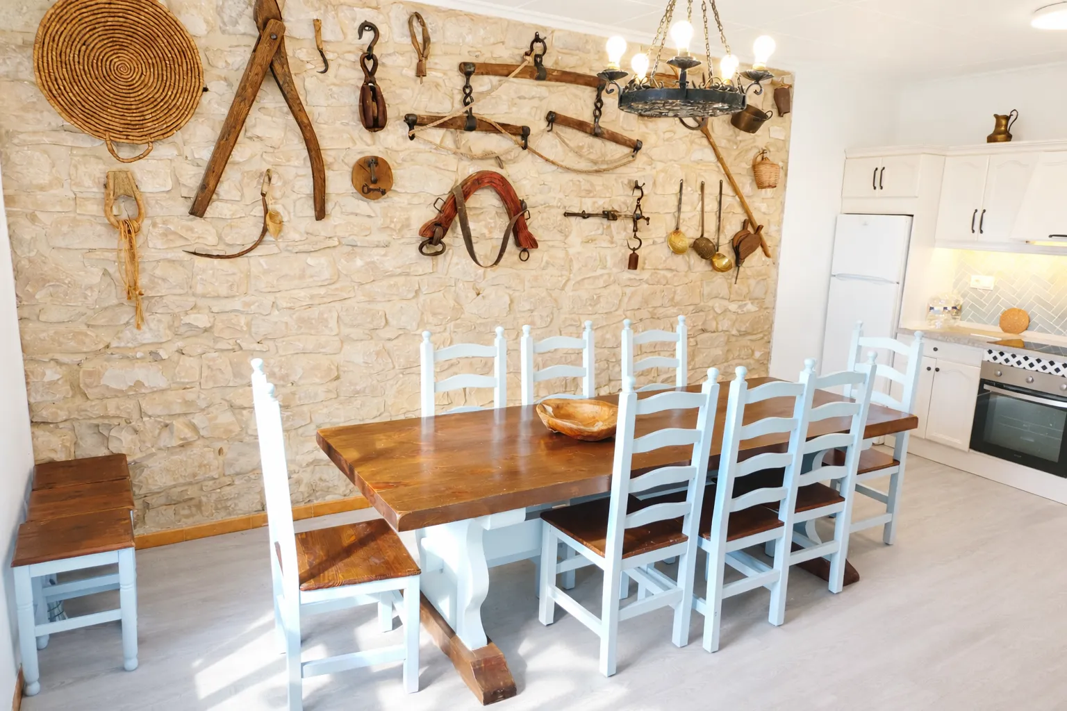 Cocina-comedor con mesa de madera, sillas azules y utensilios tradicionales en la pared de piedra